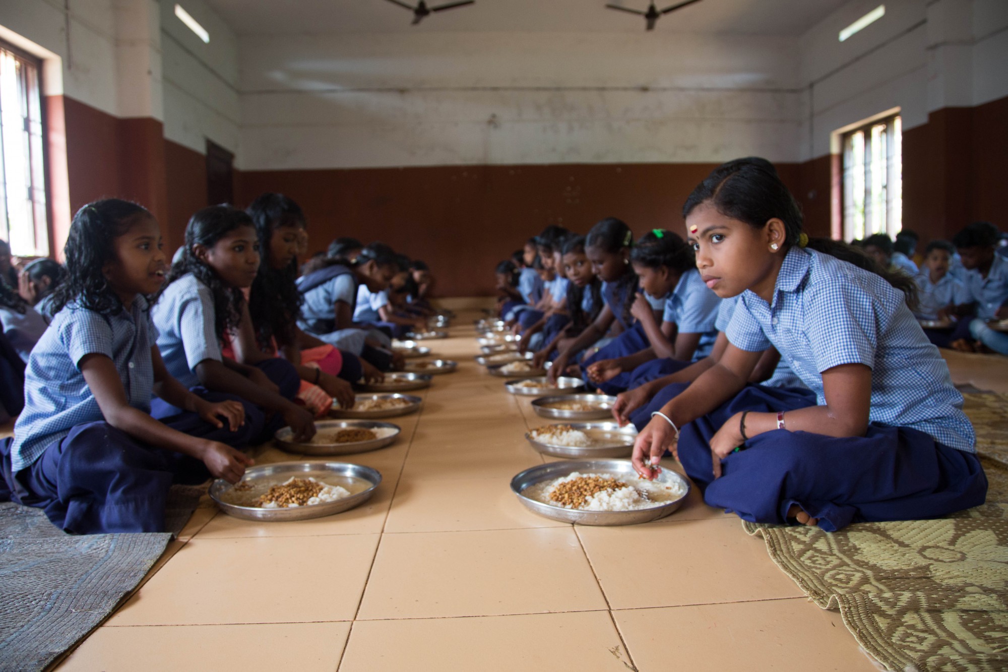 Students sit on the floor to enjoy their lunch. Some chat between mouthfuls.